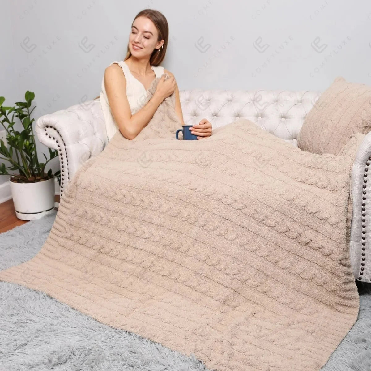 A woman sits on a white tufted sofa covered with a chunky knit beige throw blanket in a bright living room. The blanket has a textured cable-knit pattern and is draped over her and the sofa, with a gray shag rug on the floor.